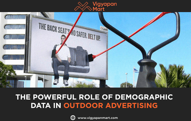 A billboard shows a man sitting on a seat, while a giant slingshot in front appears to strap him in with seatbelts. Text: THE POWERFUL ROLE OF DEMOGRAPHIC DATA IN OUTDOOR ADVERTISING. Vigyapan Mart logo is displayed above.