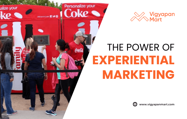 People interact with personalized Coke vending machines at an outdoor event. The image promotes the concept of experiential marketing, with the Vigyapan Mart logo and website displayed on the right.