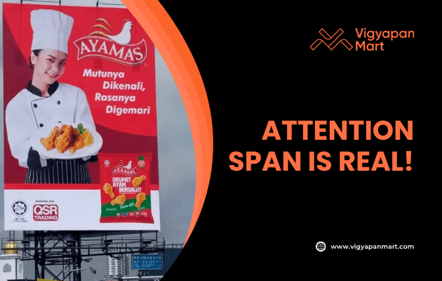 Large roadside billboard for Ayamas fried chicken featuring a smiling chef holding crispy chicken with product packaging on a red background. Overlay text reads “Attention Span is Real!” with Vigyapan Mart branding, emphasizing impactful billboard advertising.