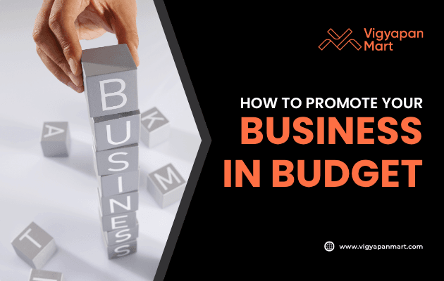 A hand stacks blocks spelling “BUSINESS” beside text that says “How to Promote Your Business in Budget,” with the Vigyapan Mart logo and website shown.