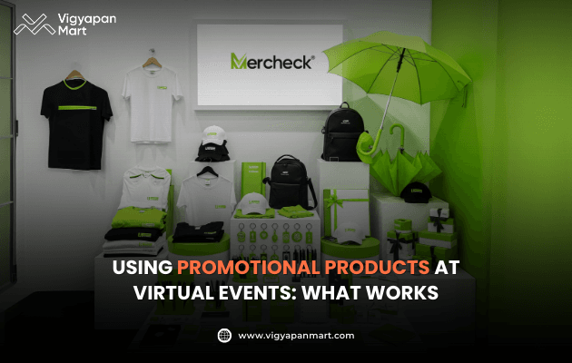 Vigyapan Mart creative showing a display of branded promotional products like t-shirts, bags, umbrellas, mugs, and gift boxes in green and white with the Mercheck logo. Text overlay reads “Using Promotional Products at Virtual Events: What Works.