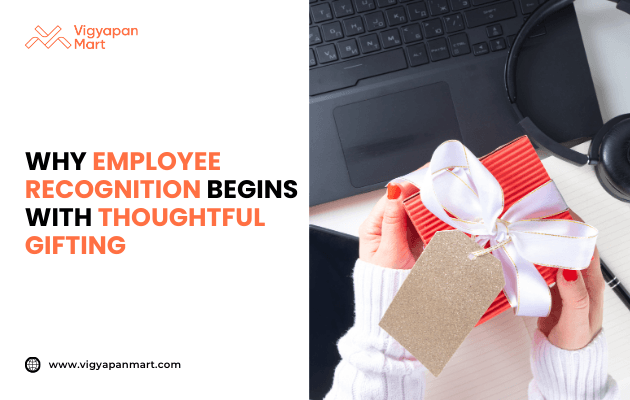 Hands holding red gift box wrapped with white satin ribbon and blank gold tag, placed near laptop and notebook on office desk. Marketing banner text reads 'Why Employee Recognition Begins with Thoughtful Gifting' by Vigyapan Mart, highlighting workplace appreciation through corporate gifts.