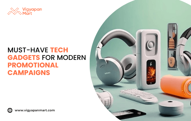 Collection of modern tech gadgets including wireless headphones, portable speakers, smart devices, and accessories displayed on a table. Text on the left reads: “Must-have tech gadgets for modern promotional campaigns” with Vigyapan Mart logo and website www.vigyapanmart.com. Tech-based corporate gifting and promotional products concept.