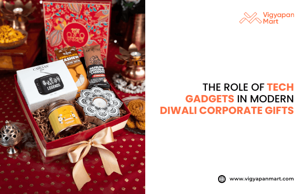 Diwali gift box with tech gadget, snacks, sweets, and golden ribbon on red cloth. Text says "The Role of Tech Gadgets in Modern Diwali Corporate Gifts." Vigyapan Mart logo top right, website bottom right. Shows mix of tech and festive gifting.