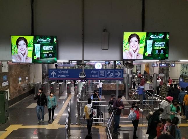 A modern Indian metro train or metro station with a clearly visible branded advertisement, showing commuters in the background.